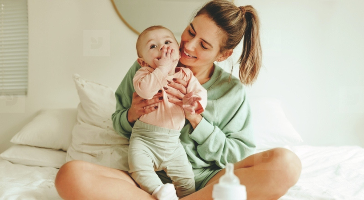 Mother in a green sweatshirt joyfully holding her baby on a bed in a softly lit bedroom.