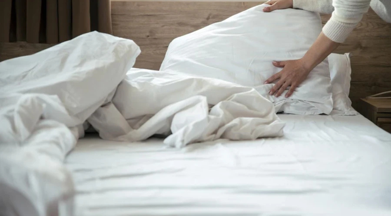 A person arranging white pillows and bedding on an unmade bed, in a cozy room with brown curtains and wooden walls.