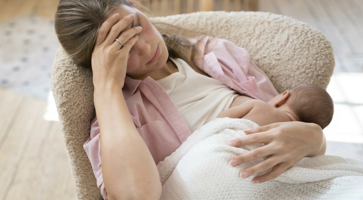 A tired mother cradles her sleeping baby while resting her head in her hand, showcasing the challenges of new motherhood.