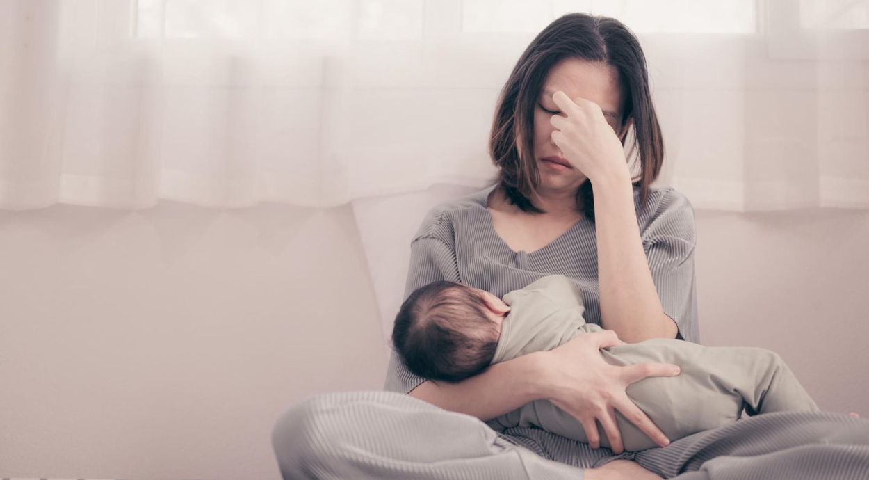 A mother gently cradles her baby while seated by a window with soft, natural light and sheer curtains.