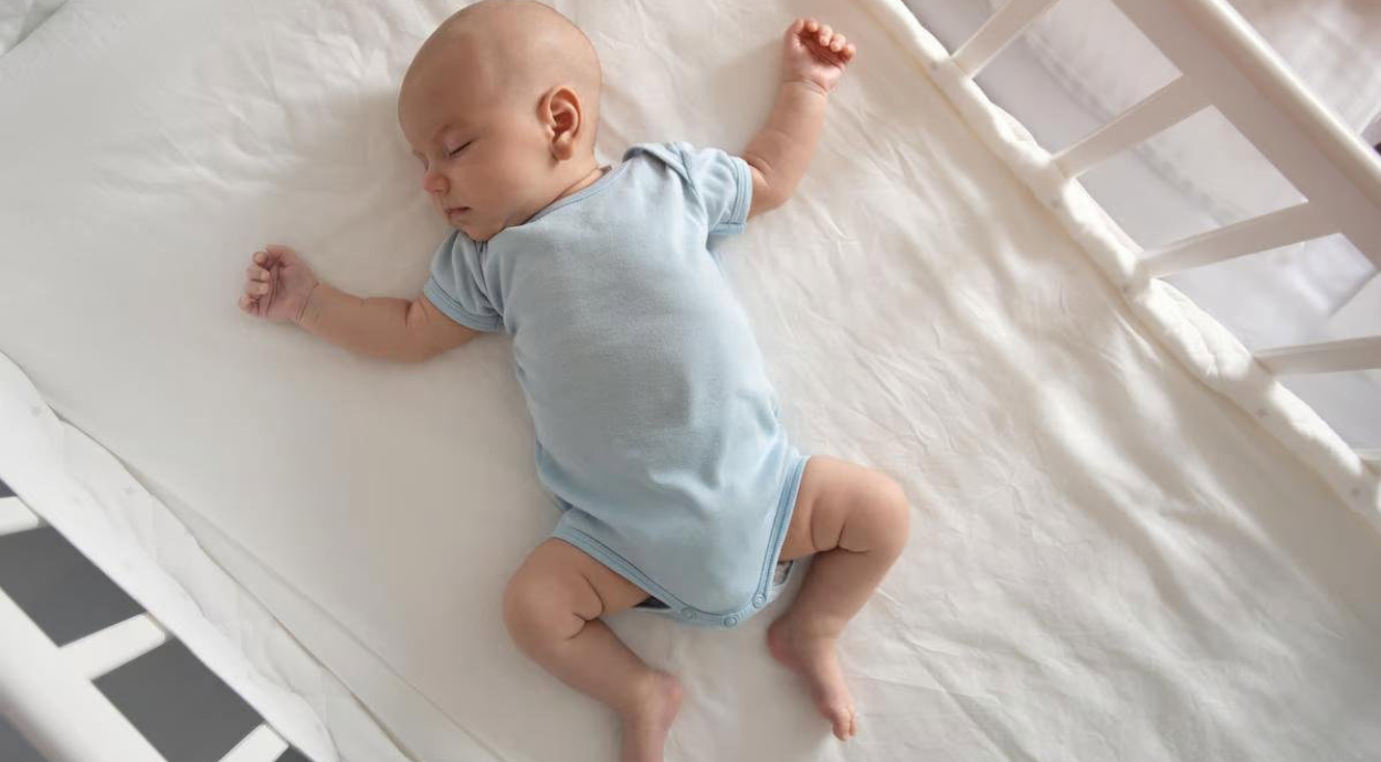 An infant in a light blue onesie is peacefully laying on a white crib mattress, arms and legs relaxed.