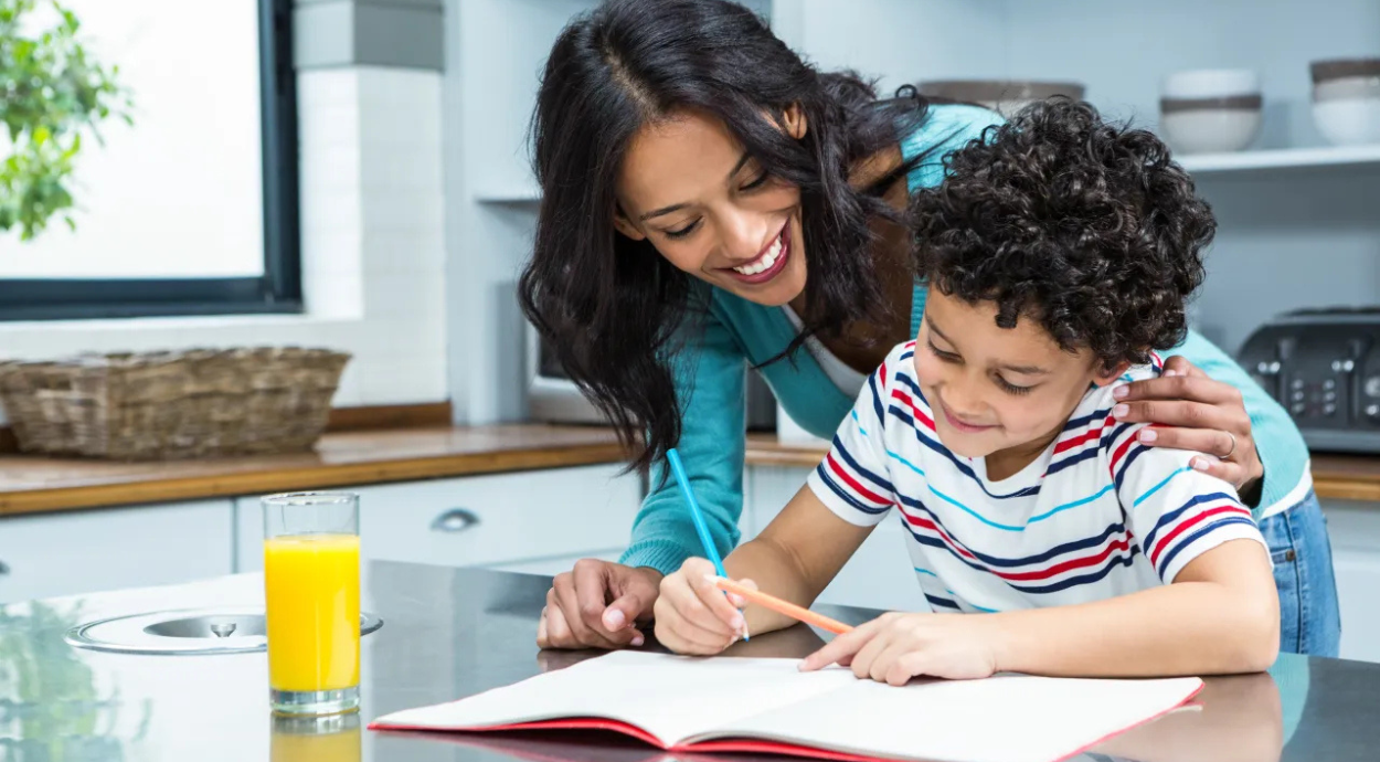 A child in a striped shirt focuses on a workbook while an adult assists, both sitting at a kitchen counter with a glass of orange juice nearby.