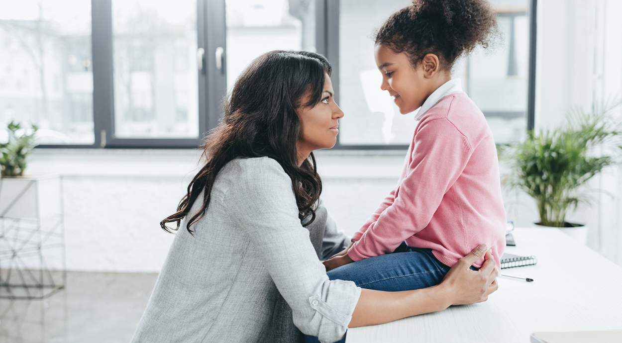 A woman kneels beside a seated girl, both engaged in a serious conversation in a well-lit, modern room with greenery.