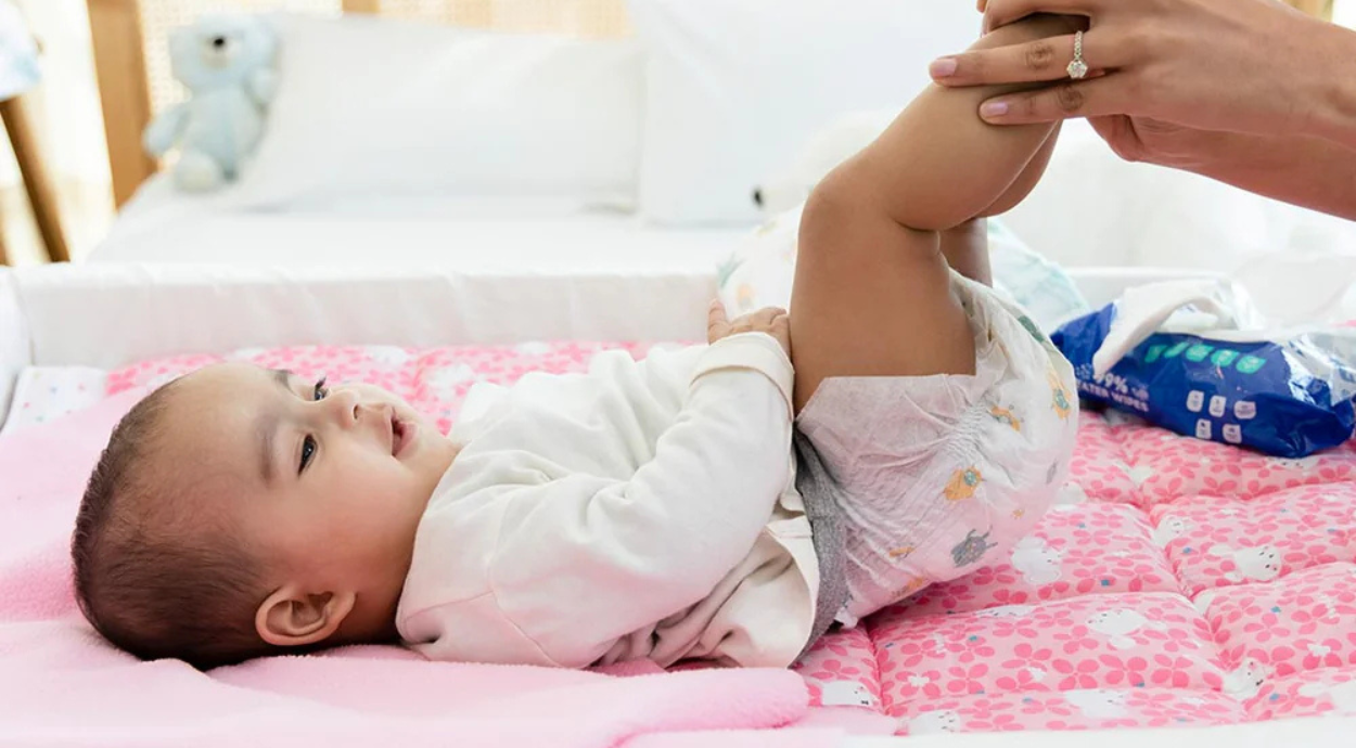 A baby lies on a pink patterned changing mat, while an adult's hand gently lifts their leg for a diaper change.