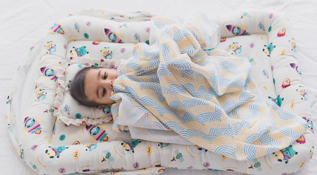 A cozy baby nest with colorful patterns, featuring soft fabrics and a decorative blanket draped over a resting baby.