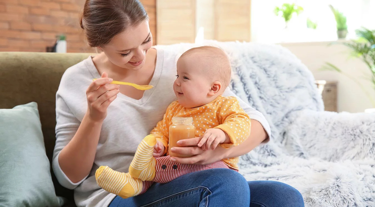 A mom sits on a couch, feeding a baby with a spoon while the child holds a jar of baby food, surrounded by soft pillows.