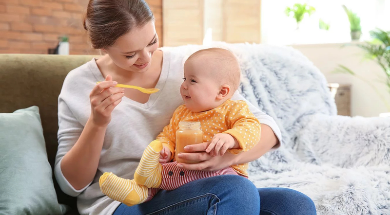 A mom sits on a couch, feeding a baby with a spoon while the child holds a jar of baby food, surrounded by soft pillows.