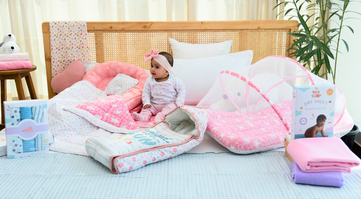 A baby wearing a bow headband sits on a bed surrounded by pink and pastel bedding, including quilts and a baby net. The scene is cozy and playful.