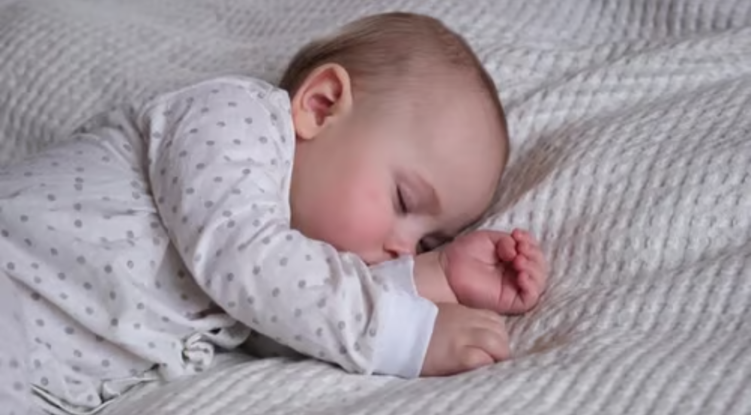Baby wearing white pajamas with gray dots sleeps curled up on a textured gray knit blanket.