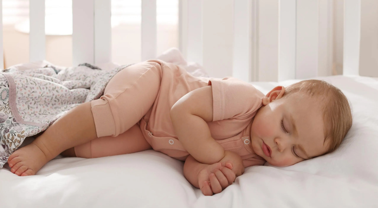 A baby sleeps peacefully on its side in a crib, wearing a soft peach onesie. The gentle lighting and pastel bedding create a calm, serene atmosphere.