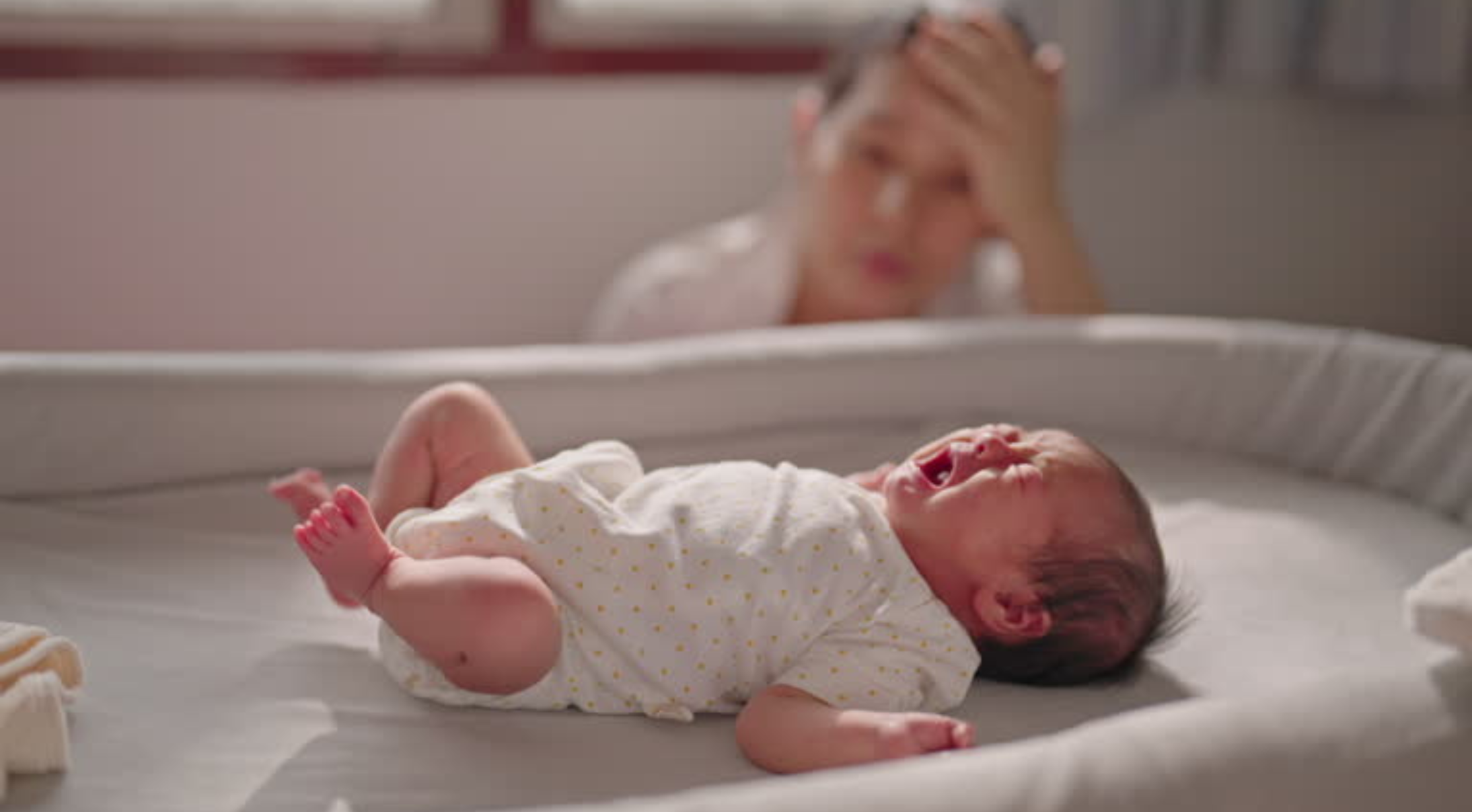 A crying baby in a light-colored onesie lies on a crib mattress, with a soft blanket nearby, in a warmly lit room.