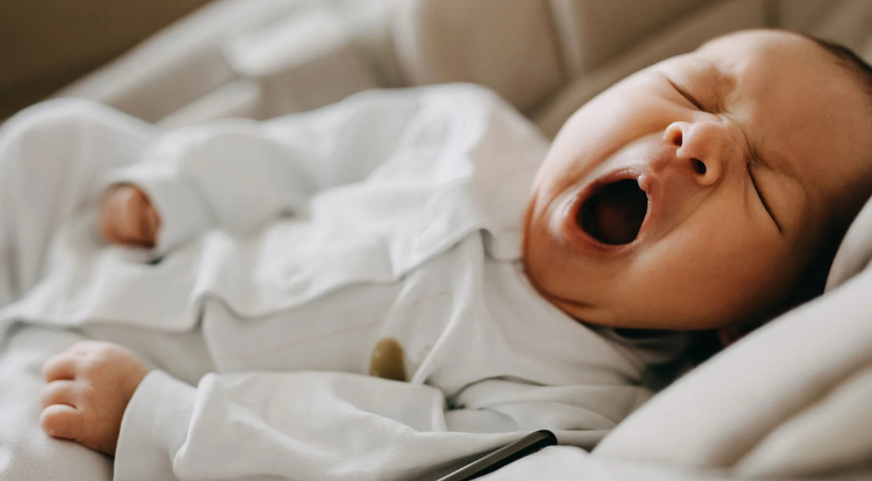 A baby is yawning widely while lying in a cozy white blanket, wearing soft white pajamas. The scene conveys a peaceful and sleepy atmosphere.