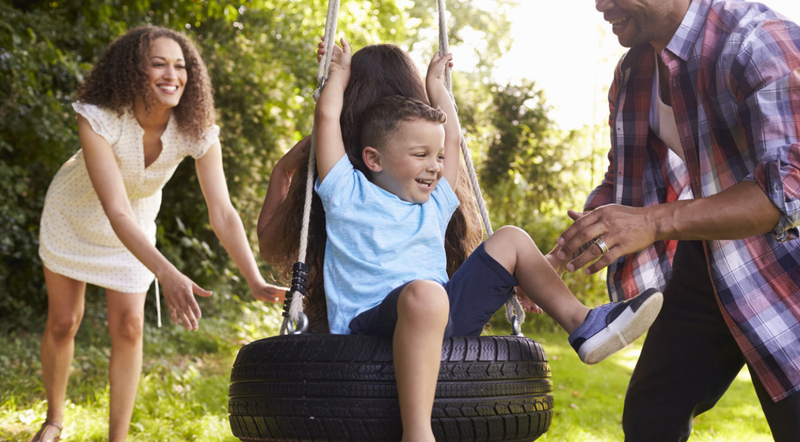 A young boy, smiling, swings on a tire swing held by a rope, with a man and woman joyfully pushing him. They're outdoors in a sunny, green garden.