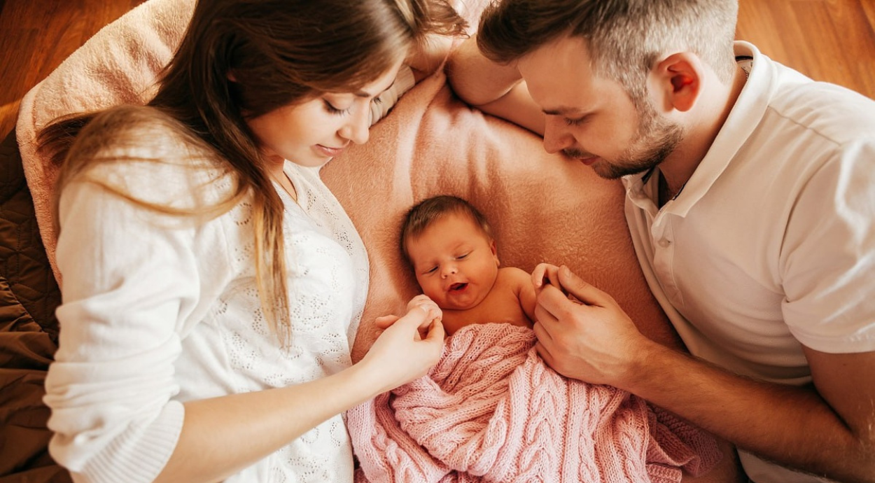 A couple lovingly cradles their newborn, wrapped in a pink blanket, on a soft blanket in a cozy setting.