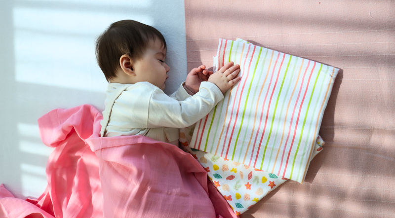 A baby peacefully sleeps on a soft surface, wrapped in a pink blanket. Nearby, colorful striped and patterned cloths add a cozy, serene atmosphere.