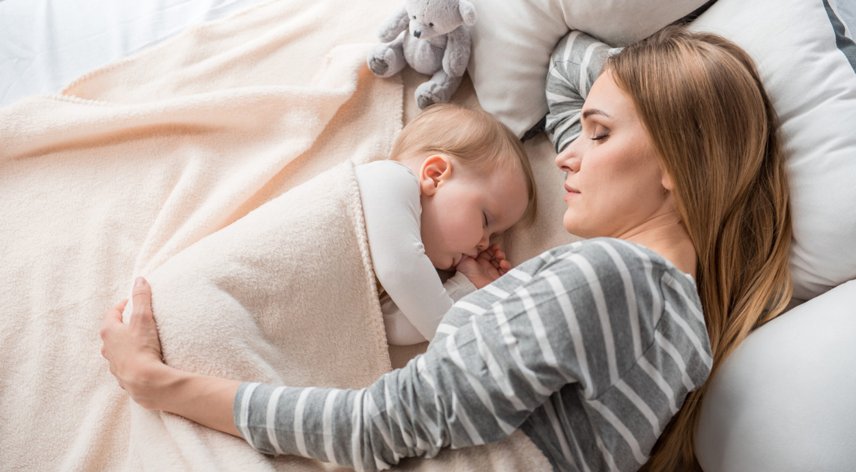A mother and baby sleep peacefully under a beige blanket. The mother, in a striped shirt, gently hugs the baby. A stuffed toy rests nearby, creating a warm, comforting scene.