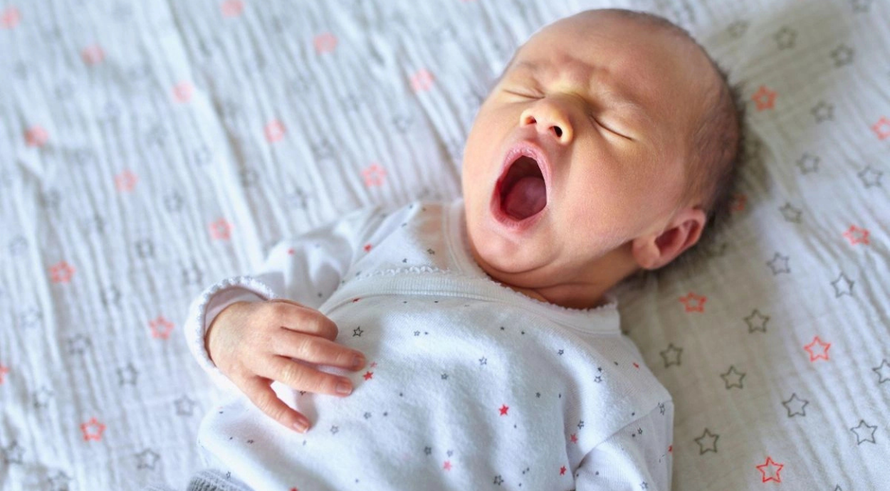 Baby wearing a white onesie with small stars yawns while lying on a patterned blanket. The scene conveys a sense of calm and sleepiness.