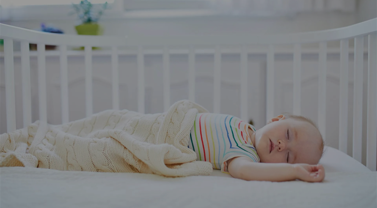A baby sleeps peacefully in a crib, wrapped in a cozy blanket, wearing a striped shirt, in a softly lit room.