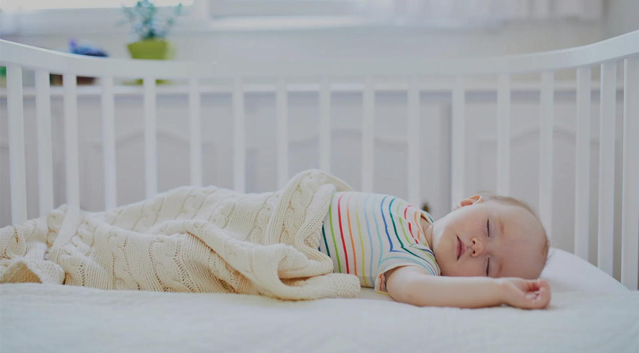 A baby peacefully asleep in a crib, wrapped in a soft beige blanket, wearing a colorful striped shirt. The room is bright and serene, creating a calm atmosphere.