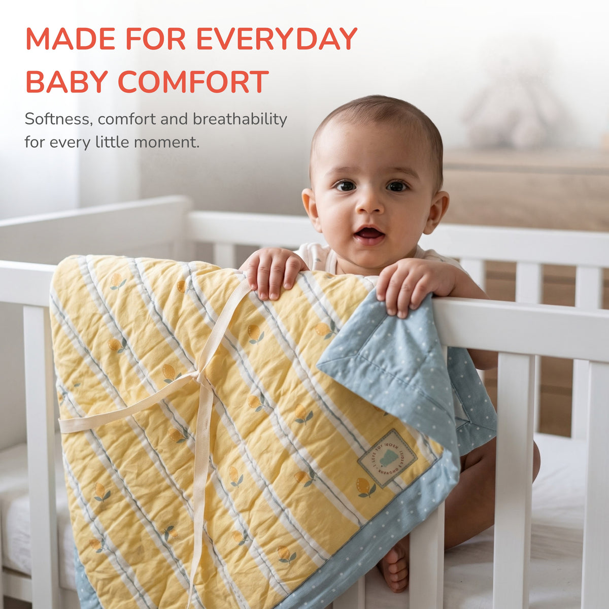 A baby sitting in a white crib holds a quilted baby blanket with a yellow and light blue design. Text at the top reads 'MADE FOR EVERYDAY BABY COMFORT' with a subtitle about softness, comfort, and breathability.