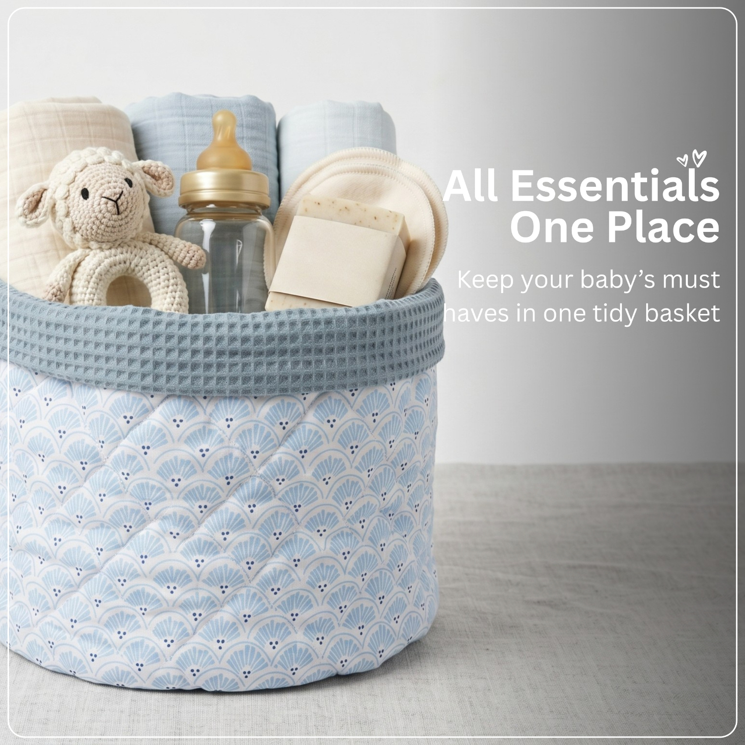 A light blue and white patterned fabric baby storage basket filled with baby essentials, including a crocheted sheep toy, a baby bottle, folded blankets, and a bar of soap. 