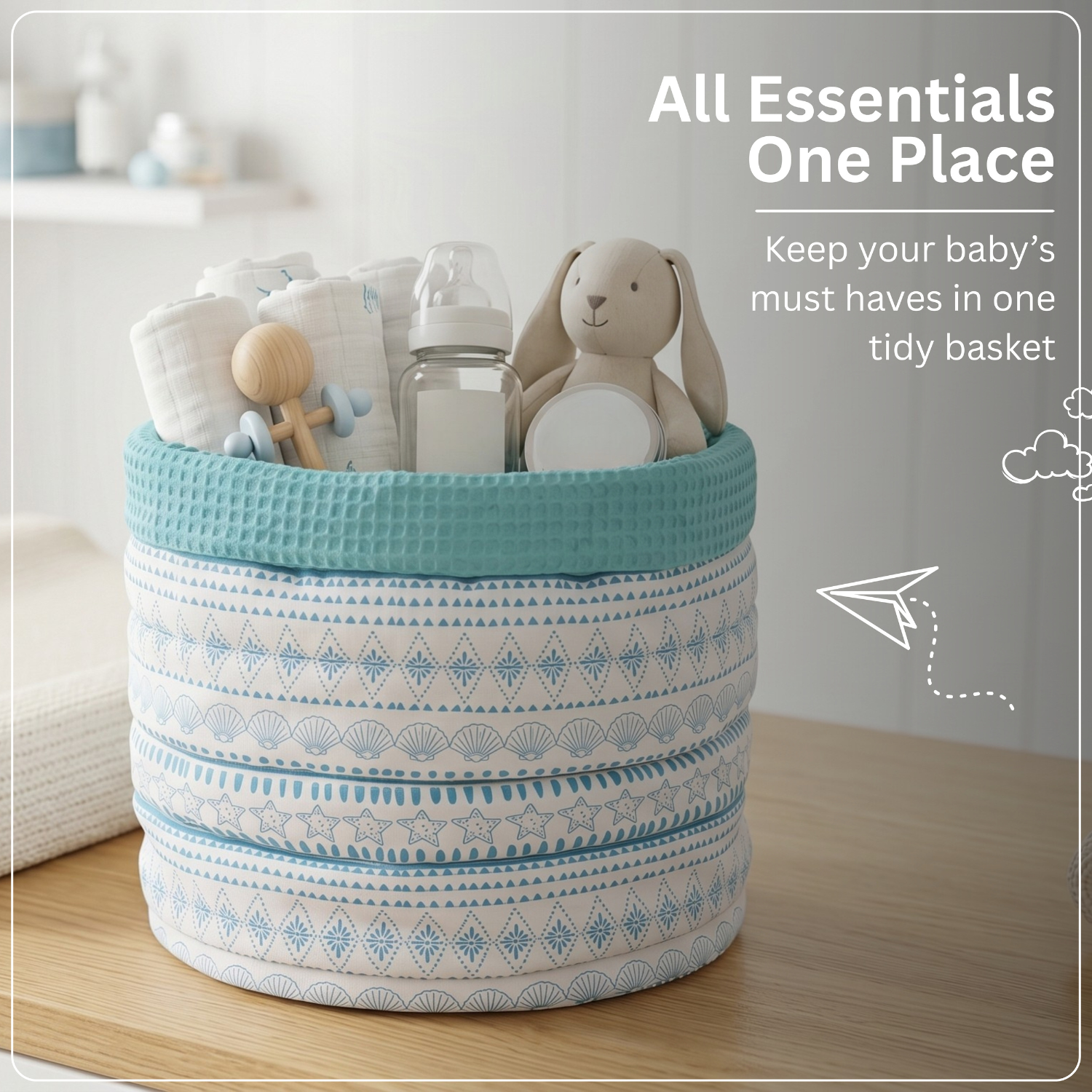 A fabric baby storage basket with a teal top rim and blue-and-white geometric patterns, filled with baby essentials including a bottle, folded cloth items, a wooden rattle, and a plush bunny.