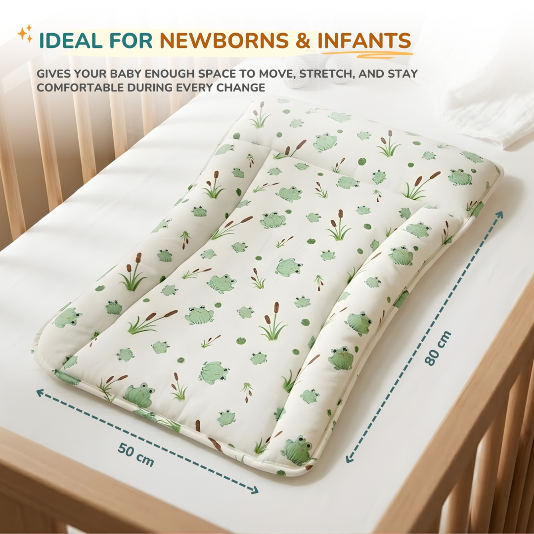 A padded baby changing station with a green frog and brown reeds print on a white background, shown on a wooden crib with text indicating dimensions of 50 cm by 80 cm and the phrase 'IDEAL FOR NEWBORNS & INFANTS'.