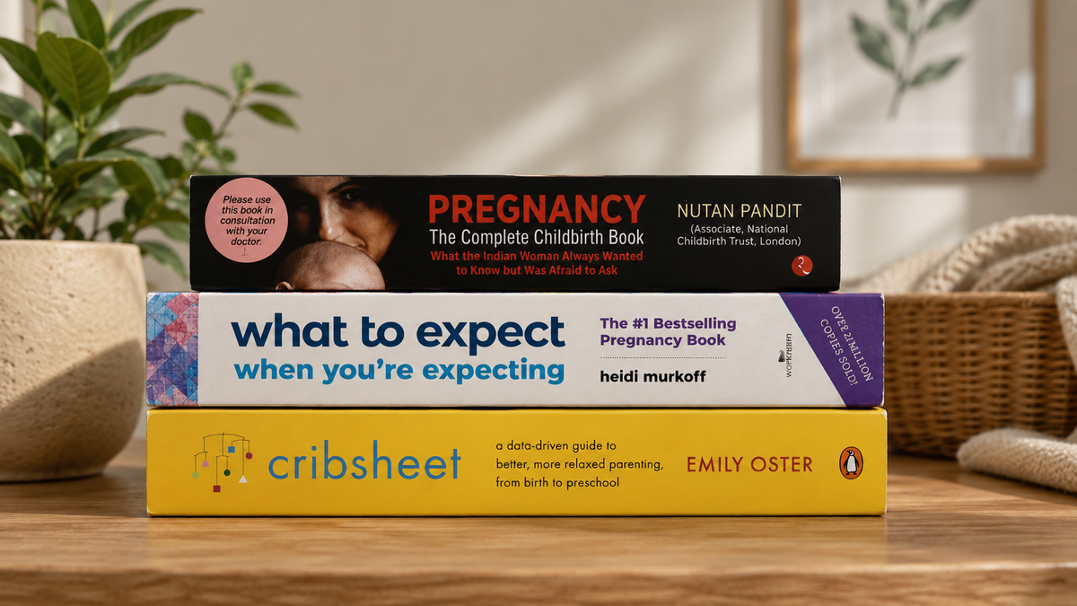 Three pregnancy and childbirth books stacked on a wooden table: 'Pregnancy: The Complete Childbirth Book' by Nutan Pandit, 'What to Expect When You're Expecting' by Heidi Murkoff, and 'Cribsheet' by Emily Oster, with a soft-focus home interior in the background.