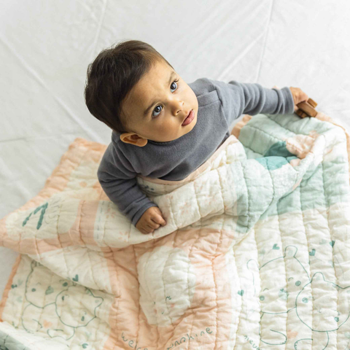 A child wrapped in a pastel-colored printed mulmul blanket, sitting on a white surface.