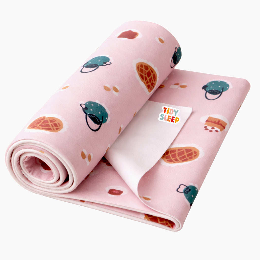 A pink baby dry sheet with a cute cartoon pattern of waffles, cappuccinos, and ice cream cones, rolled up with a white 'TIDY SLEEP' brand tag visible on the front.